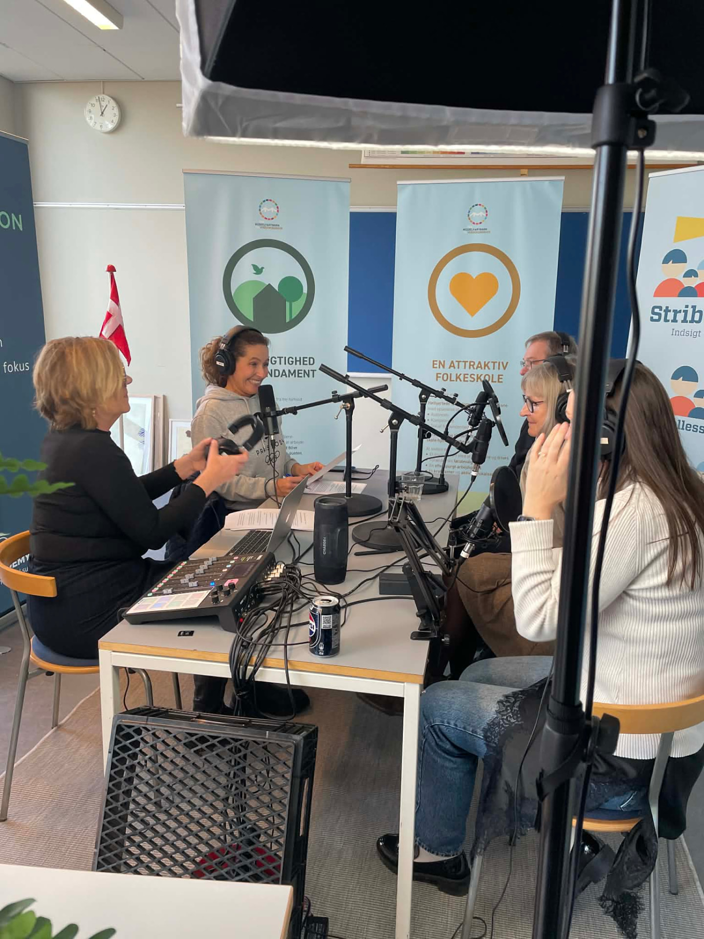 Optagelse af Strib Skoles jubilæumspodcast: fem medvirkende med høretelefoner omkring bordmikrofoner foran skolens bannere